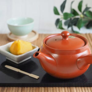 Puede incluir: Una tetera de cerámica de color terracota con tapa, boquilla y asa a juego se encuentra sobre una bandeja negra. También hay una taza de té pequeña de color verde claro y un plato cuadrado con comida amarilla. Un utensilio de madera descansa en la bandeja.