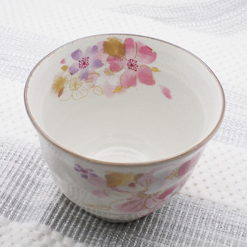 Pu&ograve; includere: Una ciotola in ceramica bianca con un motivo floreale rosa, viola e oro. I fiori sono dipinti all'esterno della ciotola. La ciotola ha una superficie leggermente strutturata e un bordo marrone.