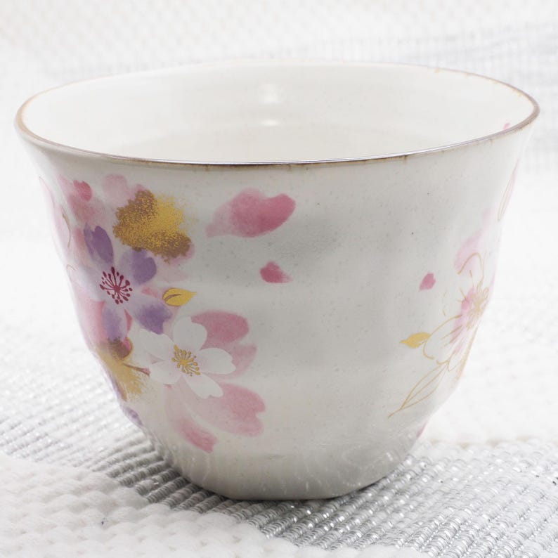 Pu&ograve; includere: Una tazza da t&egrave; in ceramica bianca con un motivo floreale. La tazza presenta illustrazioni di fiori di ciliegio rosa, viola e oro. Il bordo ha un bordo dorato. La tazza ha una superficie leggermente strutturata.