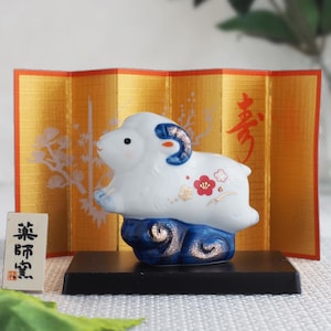 May include: A white ceramic sheep figurine with blue horns and floral accents, perched on a dark blue base. It is displayed in front of a gold folding screen with red calligraphy and white botanical illustrations, next to a small white sign with black text.