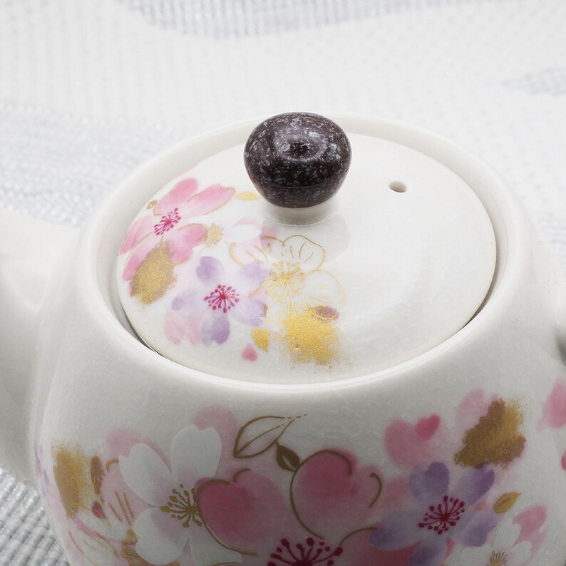 Pu&ograve; includere: Teiera in ceramica bianca con coperchio decorato con motivi floreali rosa, viola e oro. Il coperchio ha un pomello marrone scuro. La teiera &egrave; progettata per preparare il t&egrave;.