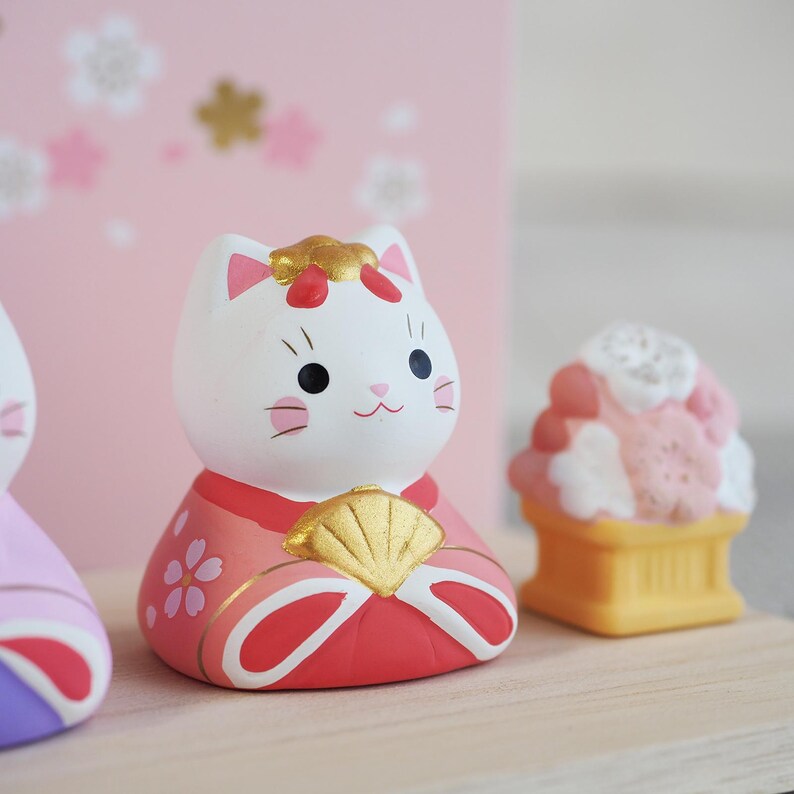 May include: A white cat figurine with a red and pink kimono, holding a gold fan. The cat has black eyes and whiskers. A pink and white dessert sits to the right. The background is pink with white floral accents.