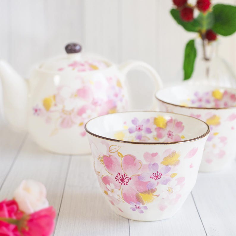 Pu&ograve; includere: Un servizio da t&egrave; in ceramica bianca con una teiera e due tazze, decorato con motivi floreali rosa, gialli e viola. Il servizio da t&egrave; ha un bordo marrone e un coperchio abbinato. Lo sfondo &egrave; una superficie di legno chiaro.