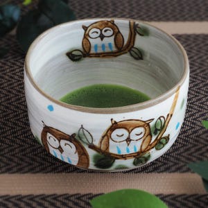 May include: A ceramic matcha bowl with a white glazed finish, featuring hand-painted brown owls perched on branches with green leaves and small blue accents. The interior contains a vibrant green tea liquid, and the rim is a light tan colour.