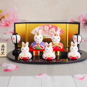 Op de afbeelding: Een decoratieve Japanse Hina-poppenset met witte konijnenfiguren met gouden accenten, gekleed in kleurrijke traditionele kleding. De set bevat een gouden scherm, een waaier en decoratieve lantaarns. De tekst op het bord luidt "薬師堂".
