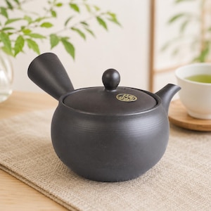 Peut inclure: Une théière kyusu en céramique noire mate avec une poignée latérale, un couvercle rond doté d'un petit bouton et un emblème circulaire doré sur le couvercle. La théière présente un bec verseur incurvé et un corps arrondi et lisse.
