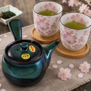以下が含まれることがあります： 濃い緑と青色のセラミックティーポットと、緑茶が入った2つのカップが特徴の日本のティーセット。カップはピンクの桜のデザインで装飾され、木製のコースターの上に置かれています。
