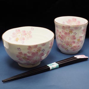 Könnte beinhalten: Ein weißes Keramik-Schalen- und Becher-Set mit rosa Kirschblüten-Designs. Das Set enthält ein Paar dunkelbraune Essstäbchen mit Kirschblüten-Design an den Spitzen. Die Schale und der Becher haben goldene Ränder.