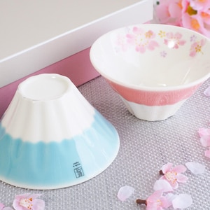 May include: Two ceramic bowls. One is white with a pink band and floral design. The other is shaped like a mountain, with a white top and blue base. The bowls are on a textured surface with pink flower petals.