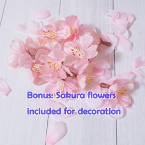 May include: A cluster of light pink artificial sakura flowers and petals on a white wooden surface. The flowers have yellow and brown centers. Text on the image reads: Bonus: Sakura flowers included for decoration.