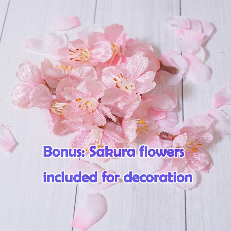 May include: A cluster of light pink artificial sakura flowers and petals on a white wooden surface. The flowers have yellow and brown centers. Text on the image reads: Bonus: Sakura flowers included for decoration.