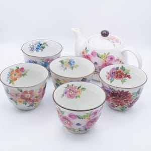 May include: A white ceramic tea set featuring a teapot and five matching bowls. Each piece is adorned with colorful floral designs in shades of pink, purple, orange, and blue. The teapot has a dark brown knob on the lid.