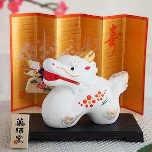 May include: A white ceramic dragon figurine with red and gold accents, holding a red stick in its mouth. The figurine has red and orange floral patterns and is set against a gold folding screen. A small wooden sign with Japanese text is in front.