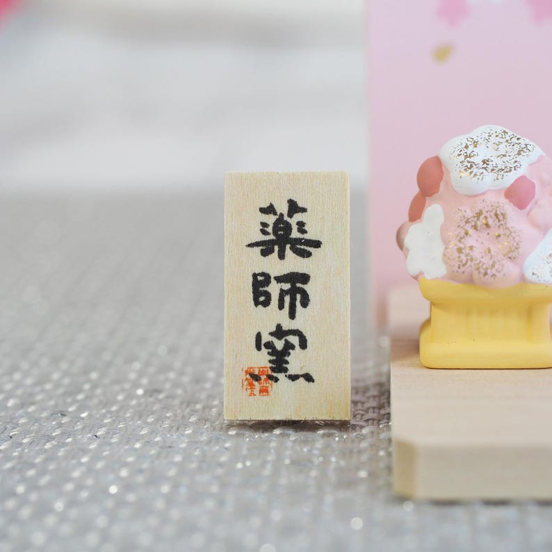 May include: A small, rectangular wooden sign with Japanese characters in black ink. The sign has a red stamp at the bottom. A colorful, decorative item is in the background.