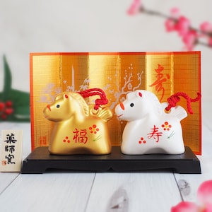 May include: Two ceramic horse figurines, one gold and one white, sit on a black stand. The gold horse has the Japanese character for "fortune" and the white horse has the character for "longevity". A gold screen is behind them.
