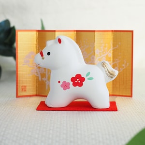 May include: A white ceramic horse figurine with red and pink floral accents. The horse has a gold-colored tail and a red ribbon around its head. It stands on a small red mat in front of a gold screen.