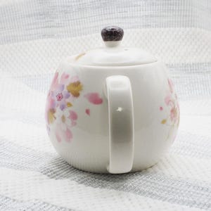 Pu&ograve; includere: Teiera in ceramica bianca con coperchio e manico. La teiera &egrave; decorata con motivi floreali rosa, oro e viola. Il coperchio ha un pomello marrone scuro. La teiera &egrave; progettata per preparare il t&egrave;.