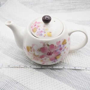 Pu&ograve; includere: Teiera in ceramica bianca con coperchio, decorata con motivi floreali rosa, viola e oro. La teiera ha un manico e un beccuccio curvi. Il coperchio &egrave; sormontato da un pomello marrone scuro. La teiera &egrave; lunga circa 18 cm.