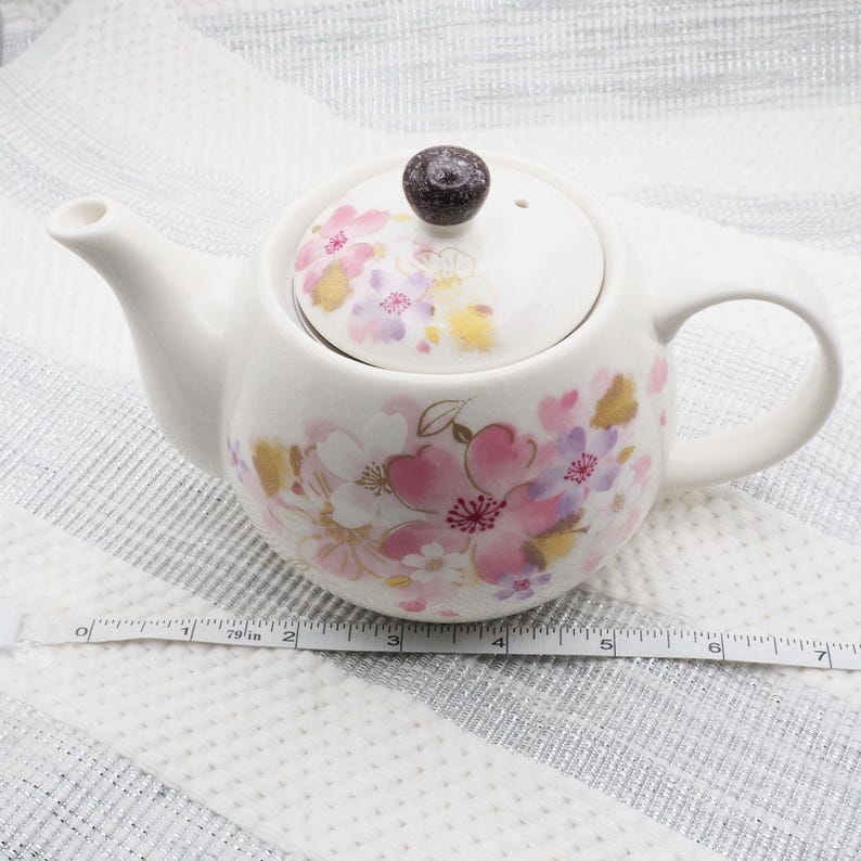 Pu&ograve; includere: Teiera in ceramica bianca con coperchio, decorata con motivi floreali rosa, viola e oro. La teiera ha un manico e un beccuccio curvi. Il coperchio &egrave; sormontato da un pomello marrone scuro. La teiera &egrave; lunga circa 18 cm.