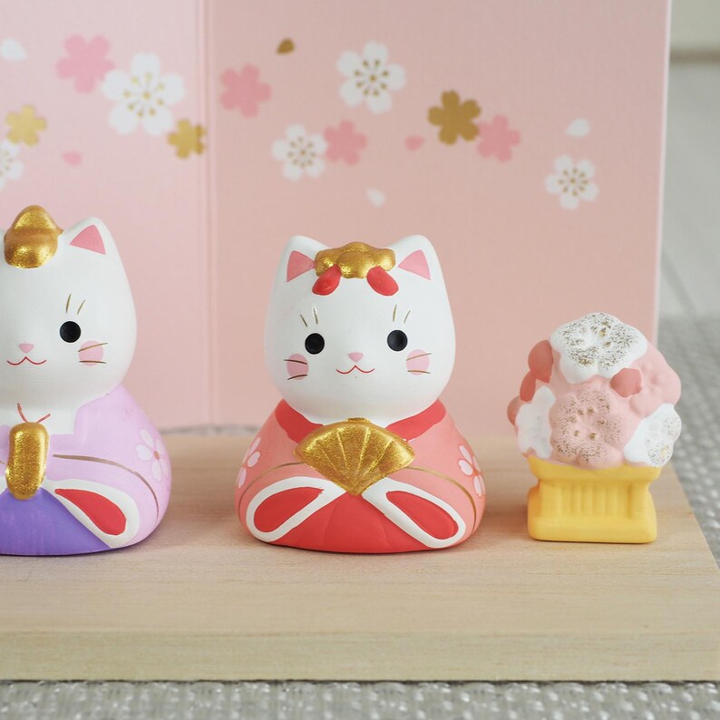 May include: Three Japanese-style cat figurines. The cats are white with gold accents and wear colorful robes. One figurine is holding a fan. A small, pink and white dessert figurine is also present. The background is pink with floral designs.