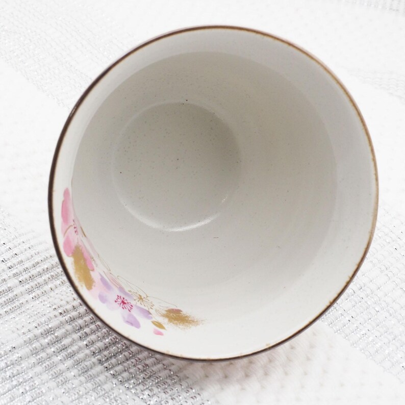 Pu&ograve; includere: Una ciotola in ceramica bianca con bordo marrone e un motivo floreale rosa, viola e oro. L'interno della ciotola &egrave; macchiettato. La ciotola &egrave; posta su uno sfondo bianco strutturato.