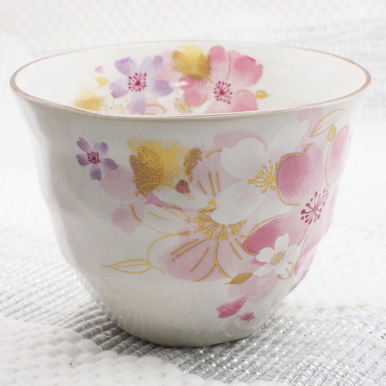 Pu&ograve; includere: Una tazza da t&egrave; in ceramica con un design floreale. La tazza presenta illustrazioni di fiori di ciliegio rosa, bianchi e dorati. La tazza ha un esterno bianco sporco strutturato e una forma leggermente irregolare.