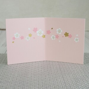 May include: A pink greeting card, open to display the inside. The card is decorated with a floral pattern of white, pink, and gold blossoms. The card is on a silver surface.
