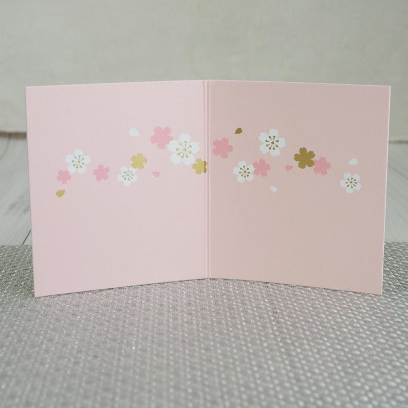 May include: A pink greeting card, open to display the inside. The card is decorated with a floral pattern of white, pink, and gold blossoms. The card is on a silver surface.