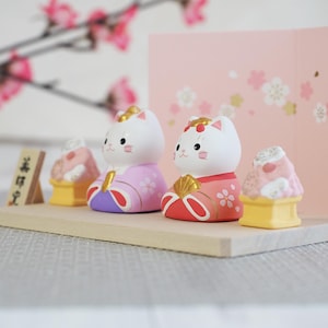 May include: A Japanese Hina Matsuri display featuring two white cat figurines in traditional attire, with gold accents and pink floral details. The display includes miniature food items and a pink backdrop with cherry blossom motifs.