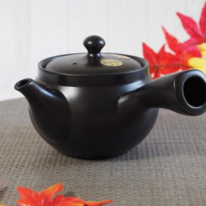 Może przedstawiać: Czarny ceramiczny czajnik z bocznym uchwytem i pokrywką z małym pokrętłem. Czajnik ma dzióbek z jednej strony i okrągły otwór z drugiej. Tło zawiera czerwone i żółte liście.