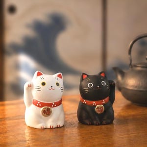以下が含まれることがあります： 白と黒の陶器製の招き猫の置物が2体。それぞれ白い水玉模様の赤い首輪と、幸運を意味する漢字が刻まれた金色の円形ペンダントを身につけ、片方の前足を上げ、顔のパーツが描かれたデザインとなっている。