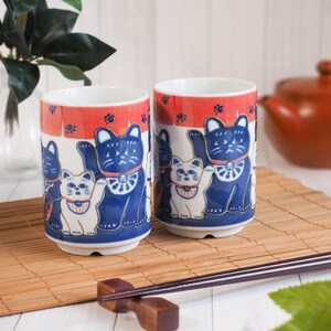 以下が含まれることがあります： 日本の招き猫のデザインが施された2つのセラミックマグカップ。マグカップは、白を基調に青と白の猫のイラスト、上部に赤い帯、足跡が特徴です。背景にはティーポットと竹のマットがあります。