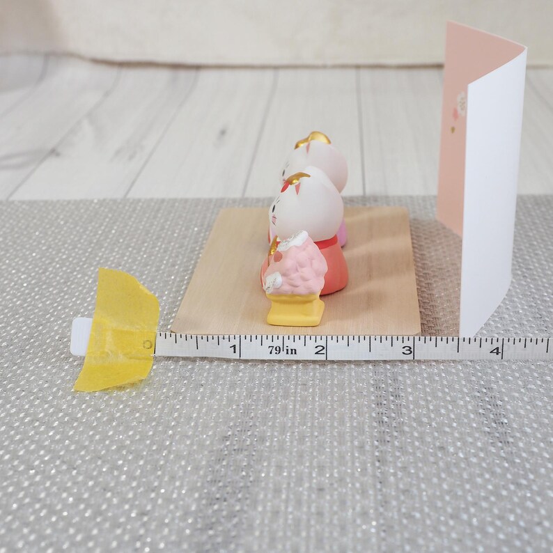 May include: A collection of Japanese Hina dolls, with a pink and white color scheme, arranged on a wooden base. A measuring tape shows the dolls are approximately 2 inches in height. A pink and white card is in the background.