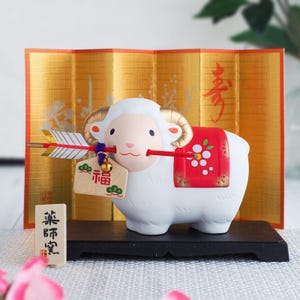 May include: A white ceramic sheep figurine with gold-colored horns, holding a red and white arrow in its mouth. A wooden tag with the red kanji character for luck hangs from the arrow. The sheep wears a red saddle with white floral patterns.