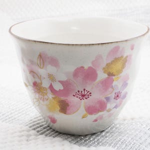 Pu&ograve; includere: Una tazza da t&egrave; in ceramica bianca con un motivo floreale. La tazza presenta illustrazioni di fiori di ciliegio rosa, bianchi e dorati. Il bordo ha un bordo dorato. La tazza &egrave; posta su uno sfondo strutturato bianco e grigio.