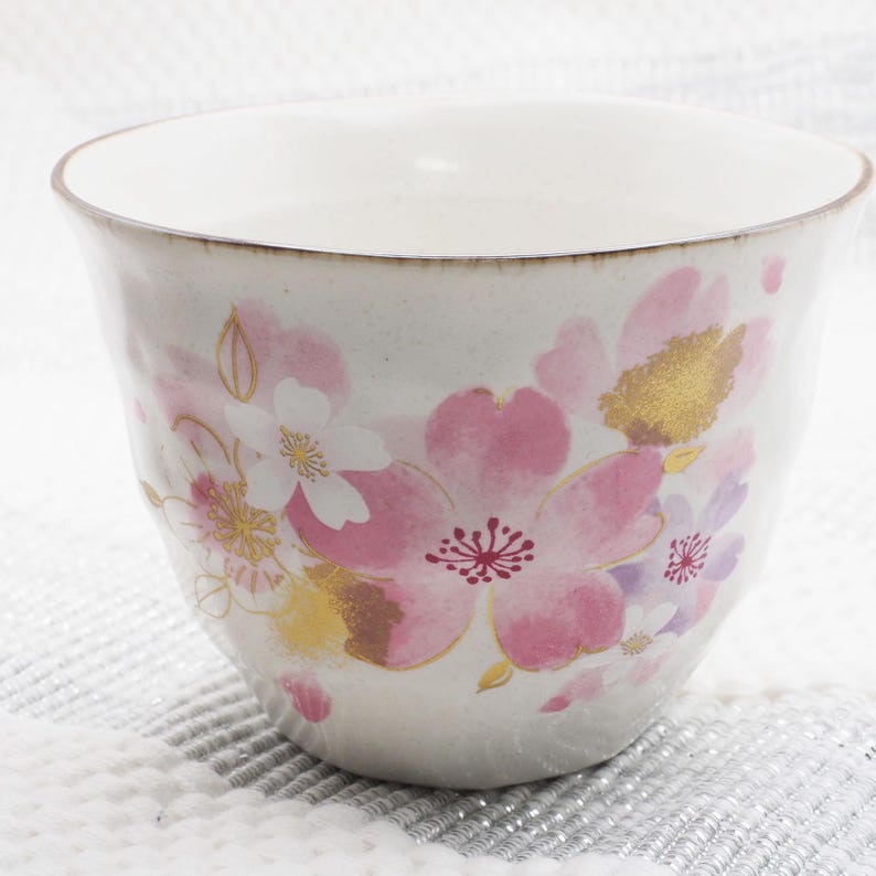 Pu&ograve; includere: Una tazza da t&egrave; in ceramica bianca con un motivo floreale. La tazza presenta illustrazioni di fiori di ciliegio rosa, bianchi e dorati. Il bordo ha un bordo dorato. La tazza &egrave; posta su uno sfondo strutturato bianco e grigio.