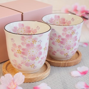 Puede incluir: Dos tazas de cerámica blanca con un estampado floral rosa, cada una sobre un pequeño posavasos de madera. Las tazas tienen un borde oscuro y están decoradas con flores de cerezo rosas. Una caja de regalo rosa está en el fondo.