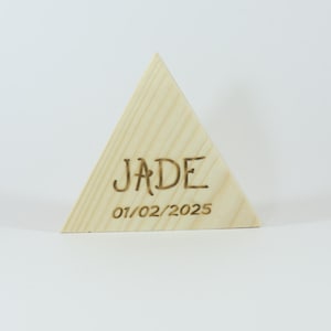 Puede incluir: Un bloque de madera triangular con el nombre "JADE" y la fecha "01/02/2025" grabados a fuego. El bloque es de color claro y se alza sobre un fondo blanco.