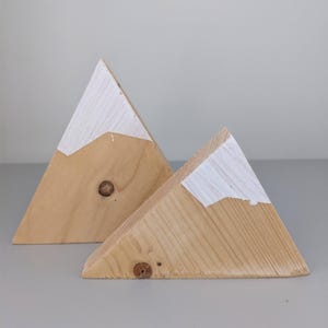 Peut inclure: Deux décorations en forme de montagne en bois. Les montagnes sont en bois clair avec un sommet peint en blanc, ressemblant à de la neige. Le bois présente des veines et des nœuds visibles. Les montagnes sont exposées sur une surface grise.