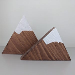 Peut inclure: Deux décorations en forme de montagne en bois. Les montagnes sont en bois brun avec des sommets peints en blanc, ressemblant à des sommets enneigés. Les formes triangulaires sont appuyées l'une contre l'autre, créant un affichage décoratif.