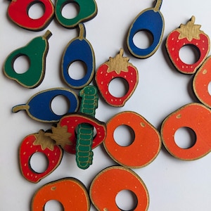 Può includere: Ritagli di legno di frutta e un verme in vari colori. Include fragole rosse, pere verdi, prugne blu e cerchi arancioni, tutti con bordi dorati. È presente anche un ritaglio di verme verde. Probabilmente per un giocattolo o un progetto artigianale.