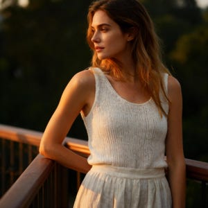 May include: A woman wearing a white vest top and a cream-coloured skirt, standing on a wooden deck. The vest top has a scoop neck and a ribbed texture. The skirt is a flowing, pleated design. The background is a blurred view of trees.