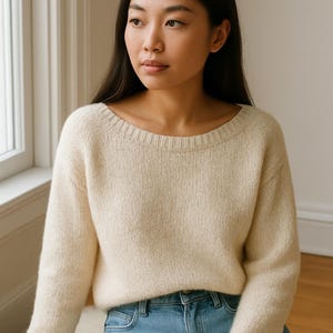 May include: A cream-coloured sweater with a wide neckline and long sleeves. The sweater is paired with light blue denim jeans. The model is sitting near a window, with a neutral background.