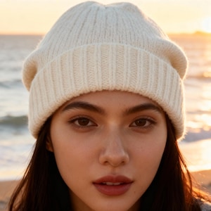 Può includere: Berretto lavorato a maglia color crema. Il cappello è realizzato in materiale a maglia a coste e ha un bordo ripiegato. Il cappello è indossato da una persona con capelli scuri e occhi marroni. Lo sfondo è una scena di spiaggia sfocata con un tramonto.