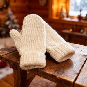 May include: A pair of ivory-colored knitted mittens rests on a wooden surface. The mittens have a ribbed cuff and a thick, textured knit. The background features a cozy, rustic interior with a Christmas tree and a window overlooking a snowy landscape.