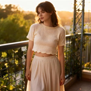 Può includere: Top corto in maglia color crema con maniche corte e orlo smerlato. Abbinato a una gonna a vita alta abbinata. L'outfit è indossato all'aperto, con uno sfondo sfocato di verde e luce solare.