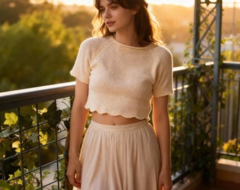 May include: Cream-colored knit crop top with short sleeves and a scalloped hem. Paired with a matching high-waisted skirt. The outfit is worn outdoors, with a blurred background of greenery and sunlight.