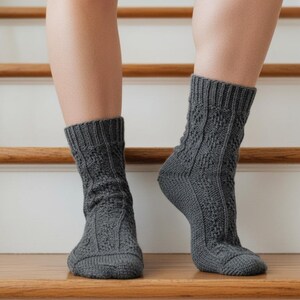 May include: A pair of gray knitted socks with a textured pattern. The socks are worn on a person's feet, with the legs visible from the ankles down. The socks are on a wooden surface with a white background.