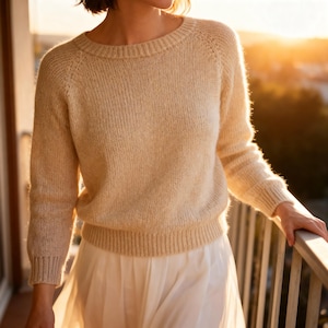 May include: A beige knit sweater with a round neckline and long sleeves. The sweater has a ribbed hem and cuffs. The model is wearing a white skirt. The background is blurred, with a warm, golden light.