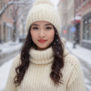 May include: A person wearing a cream-coloured knitted hat with a bobble and a matching roll-neck sweater. The person has dark hair and is smiling. Snow is falling in the background, creating a winter scene.